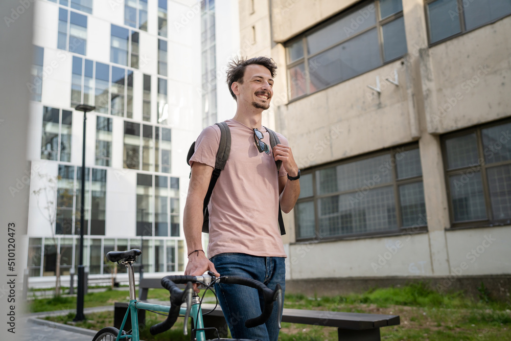 One man young adult male with brown hair and mustaches walking by the building with rucksack on his back and bicycle happy smile joyful real people copy space side view