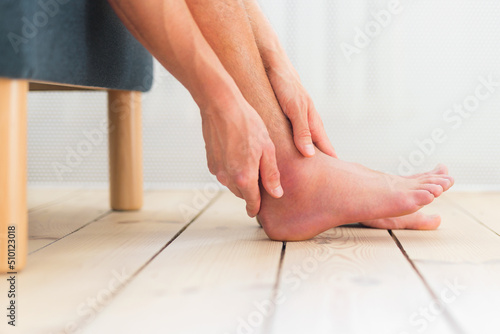 Fototapeta Foot pain, man hold his ankle sitting on the sofa at home
