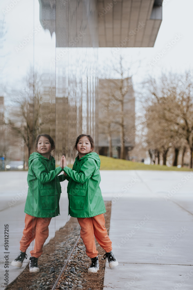 Young girl in a green raincoat dances with her reflection in a window. Young girl in a green raincoat dances with her reflection in a window.