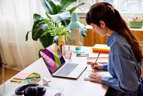 Woman writing in notepad while working on laptop photo