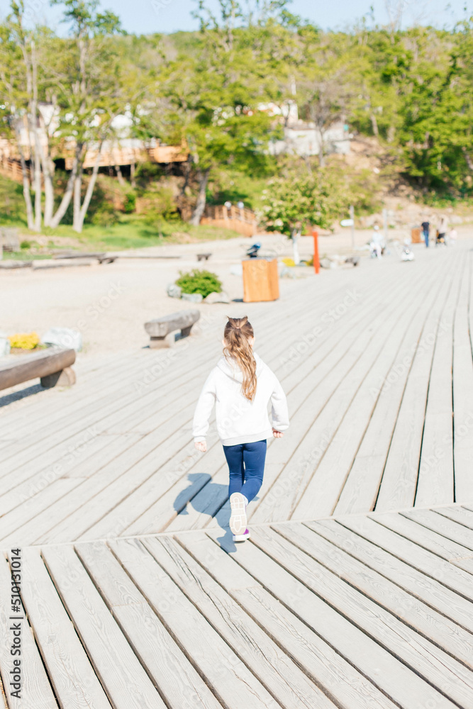 A little girl runs along a wooden path by the sea. View from the back. Coast