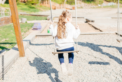 A little girl swings on a wooden swing in the park on the playground. Back view. Outdoor activities photo