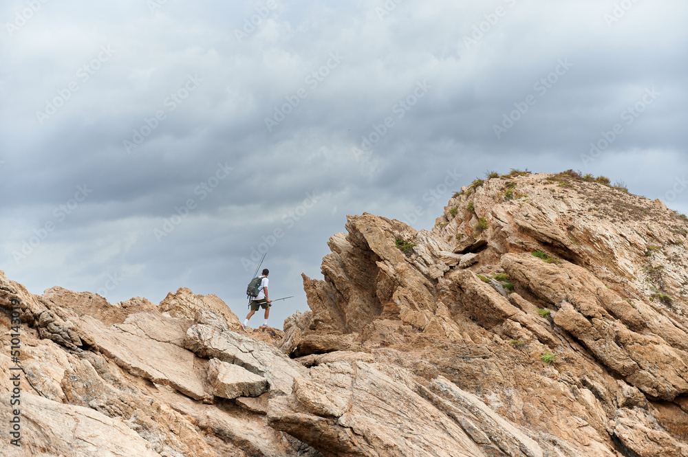 Young man carrying a backpack with sport fishing equipment goes to a fishing point hiking in Rodos Islet, Portman, Murcia coast. Spinning mode fisherman. Young man carrying a backpack with sport fishing equipment goes to a fishing point hiking in Rodos Islet, Portman, Murcia coast. Spinning mode fisherman.