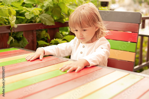 a little blond boy in a white linen shirt is sitting at a table in an outdoor cafe in the rays of the setting sun