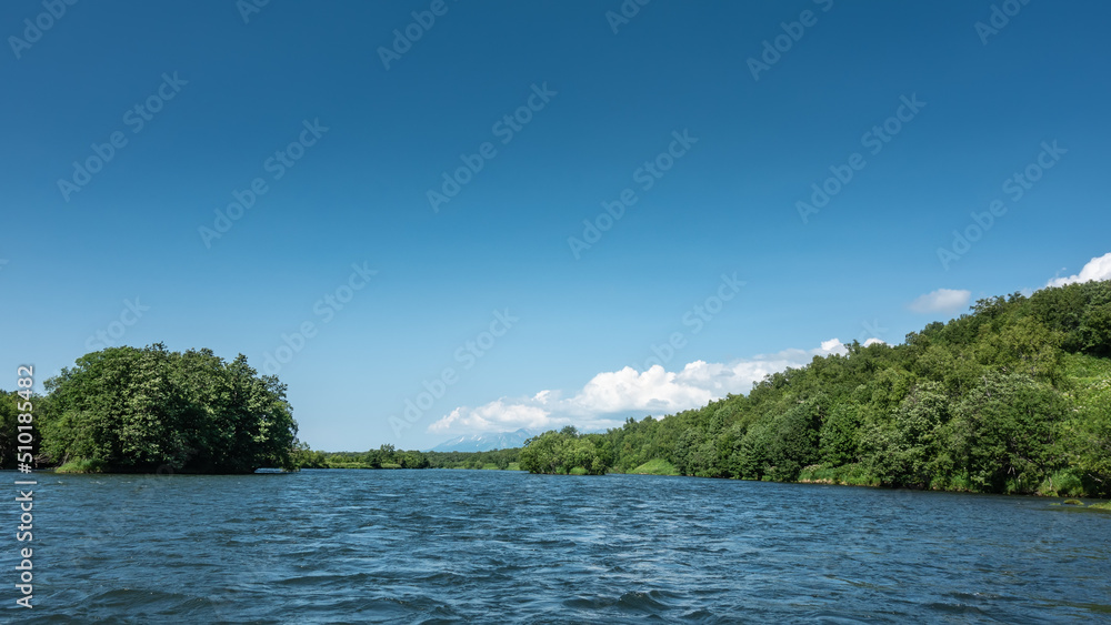 A calm blue river under an azure sky. Lush green vegetation on hilly shores. White clouds. Copy space. Kamchatka. River Bystraya