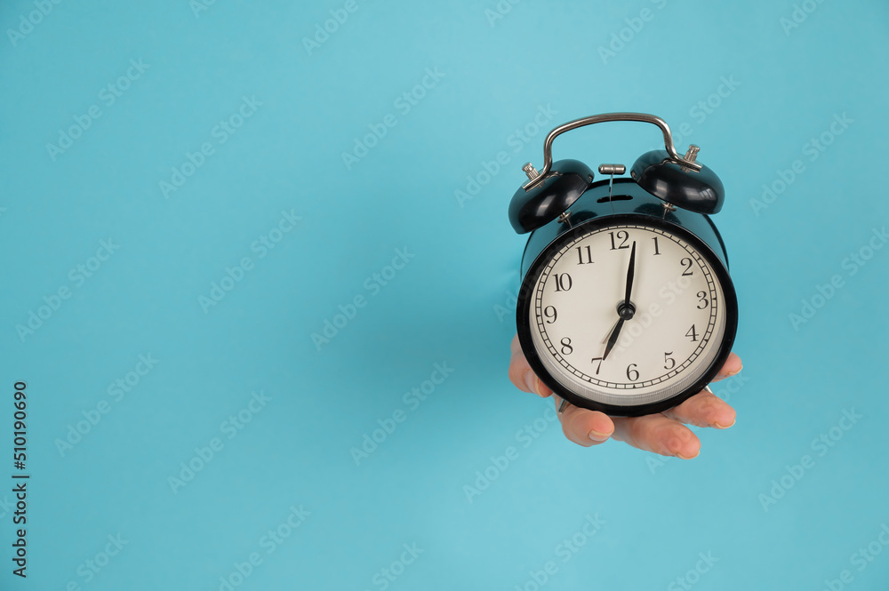 A woman's hand sticks out of a hole in a blue paper background and holds an alarm clock. Copy space. A woman's hand sticks out of a hole in a blue paper background and holds an alarm clock. Copy space.