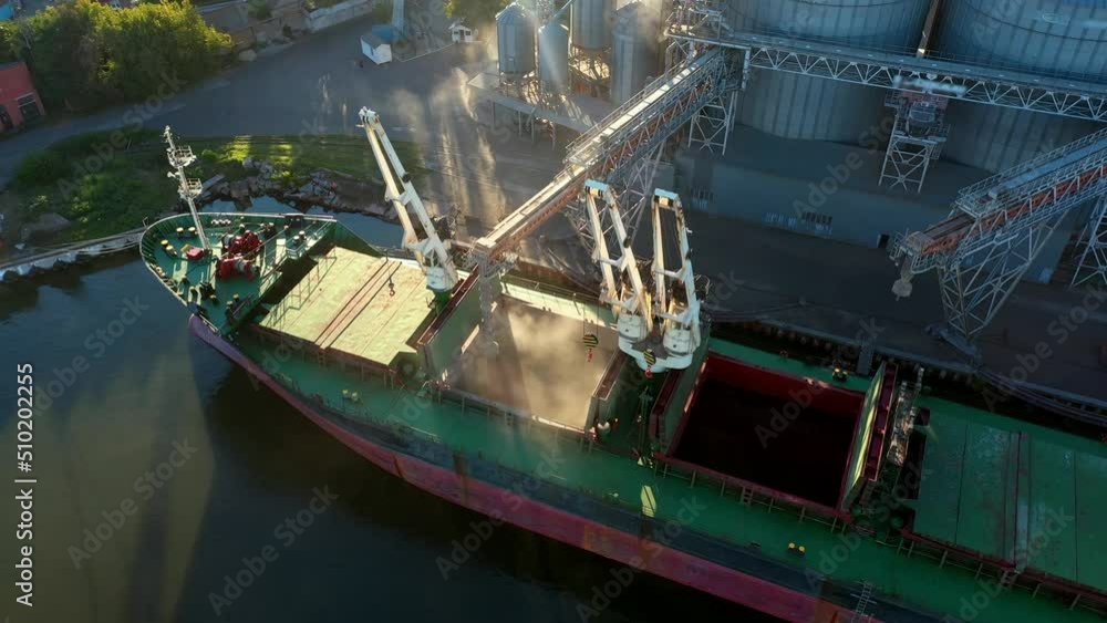 Aerial of wheat loading to bulker ship cargo hold at sea grain elevator in sea port. Wheat shipment from silo to bulk vessel via trunk at sea port terminal. Transportation of agricultural commodities. Aerial of wheat loading to bulker ship cargo hold at sea grain elevator in sea port. Wheat shipment from silo to bulk vessel via trunk at sea port terminal. Transportation of agricultural commodities.