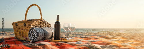 Blanket with picnic basket, bottle of wine and glasses on sandy beach near sea, space for text. Banner design