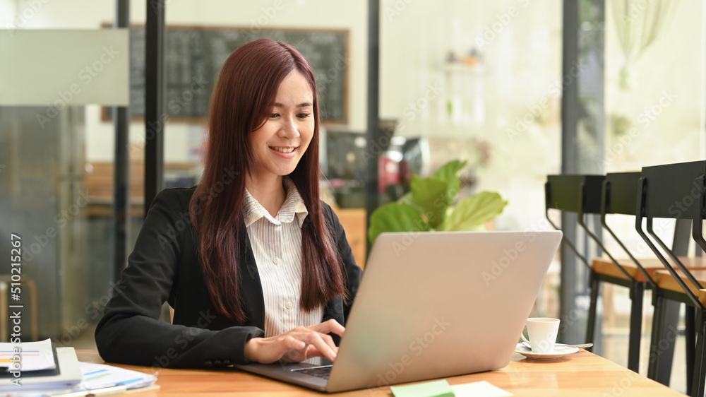 Smiling asian woman in business suit using laptop computer at coffee shop Smiling asian woman in business suit using laptop computer at coffee shop