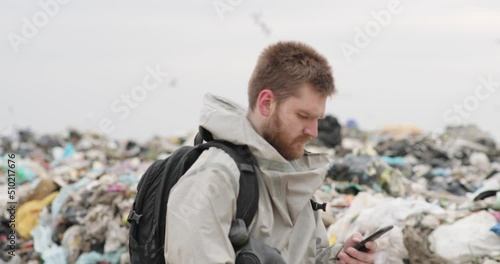 A working man against the backdrop of burning garbage. A lot of plastic bags thrown to the dump. From the plastic debris goes smoke. On the man, the khaki color encephalic suit photo