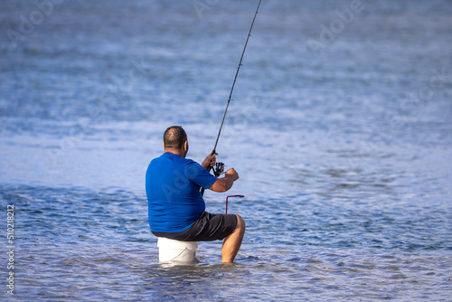 An unidentifiable middle aged man with a fishing rod sits on a bucket in a shallow lake, surrounded by calm blue water, on a sunny afternoon. Copy space.