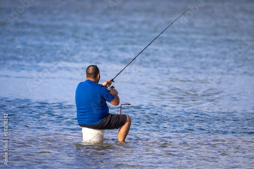 An unidentifiable middle aged man with a fishing rod sits on a bucket in a shallow lake, surrounded by calm blue water, on a sunny afternoon. Copy space.