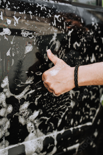 A male professional worker, a washerman is satisfied, satisfied with his work, showing thumbs up against the background of a black surface of a car door and white soap suds. Photo, copy space.