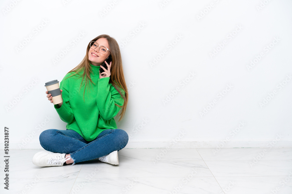 Young caucasian woman sitting on the floor isolated on white background holding coffee to take away and a mobile Young caucasian woman sitting on the floor isolated on white background holding coffee to take away and a mobile
