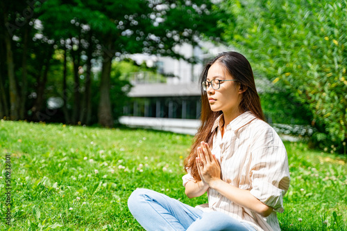 Young Asian Thai, Vietnamese or Chinese woman in casual clothes sitting in lotus yoga pose meditating on green grass loan near trees and bushes on weekend, vacation or holiday on sunny summer day. 