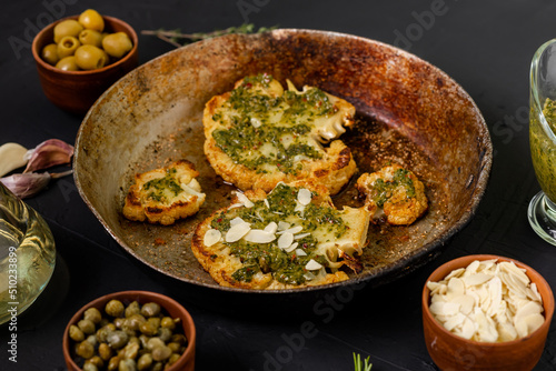 Cauliflower steak with spices lies in a frying pan. Olive oil, chimichurri sauce, capers, olives, herbs, various spices side by side. Dark background. Vegetarian food.