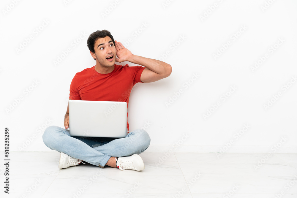 Caucasian handsome man with a laptop sitting on the floor listening to something by putting hand on the ear Caucasian handsome man with a laptop sitting on the floor listening to something by putting hand on the ear