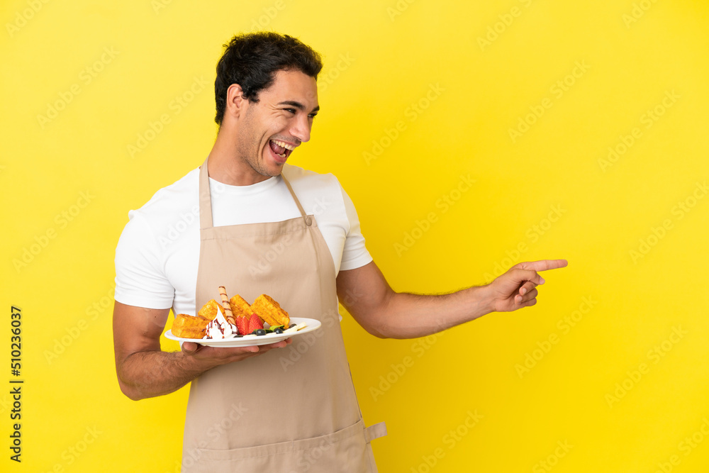 Restaurant waiter holding waffles over isolated yellow background pointing finger to the side and presenting a product