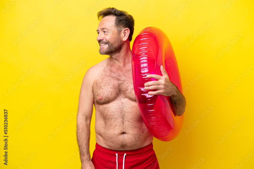 Middle age caucasian man holding inflatable donut isolated on yellow background looking to the side and smiling