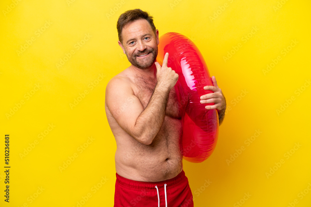 Middle age caucasian man holding inflatable donut isolated on yellow background pointing back