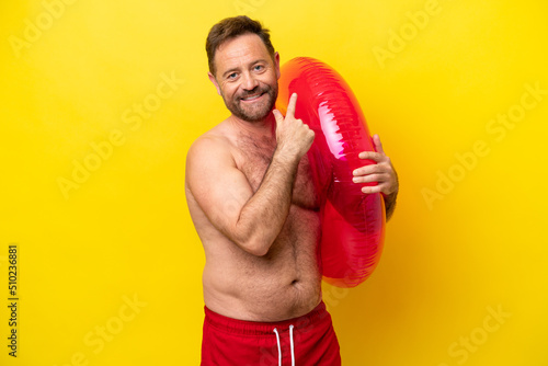 Middle age caucasian man holding inflatable donut isolated on yellow background pointing back