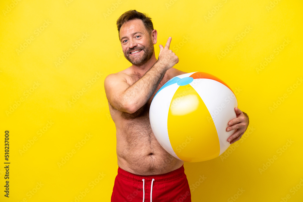 Middle age caucasian man holding beach ball isolated on yellow background pointing back