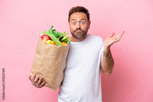 Middle age man holding a grocery shopping bag isolated on pink background having doubts while raising hands
