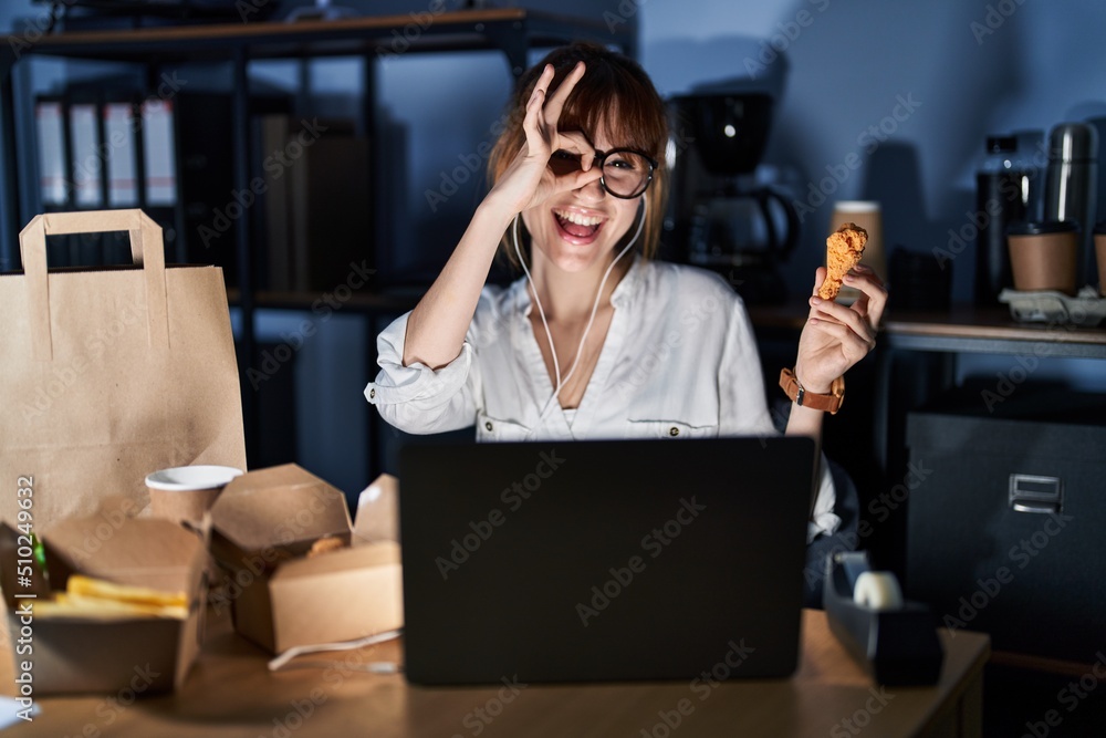 Obraz premium Young beautiful woman working using computer laptop and eating delivery food doing ok gesture with hand smiling, eye looking through fingers with happy face.