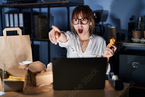 Young beautiful woman working using computer laptop and eating delivery food pointing with finger surprised ahead, open mouth amazed expression, something on the front