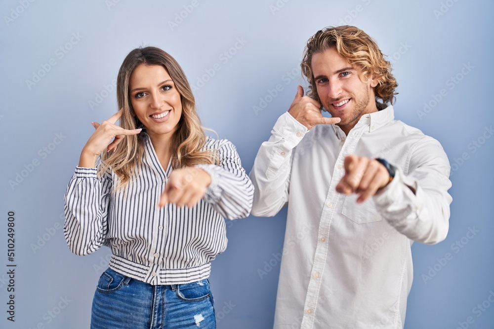 Young couple standing over blue background smiling doing talking on the telephone gesture and pointing to you. call me. Young couple standing over blue background smiling doing talking on the telephone gesture and pointing to you. call me.