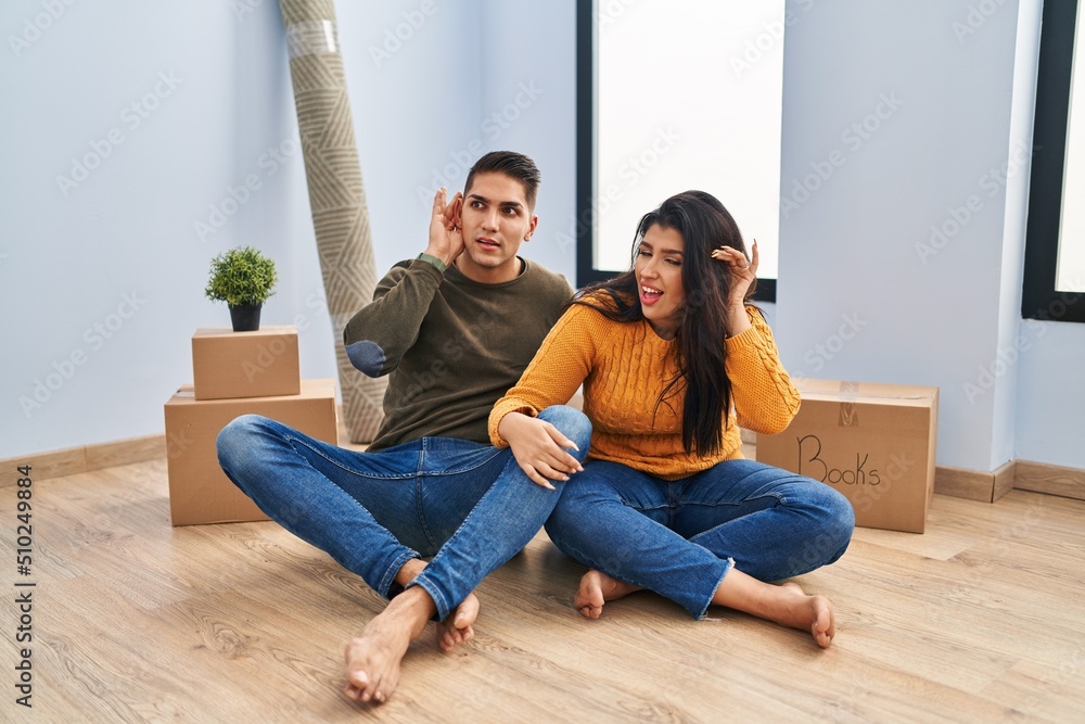 Young couple sitting on the floor at new home smiling with hand over ear listening an hearing to rumor or gossip. deafness concept. Young couple sitting on the floor at new home smiling with hand over ear listening an hearing to rumor or gossip. deafness concept.