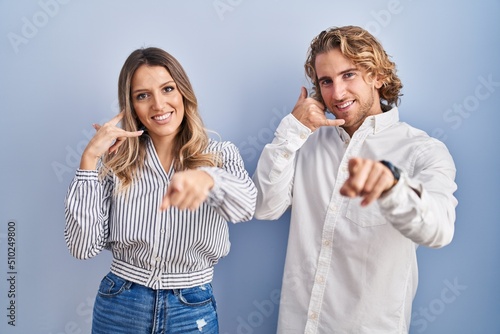 Young couple standing over blue background smiling doing talking on the telephone gesture and pointing to you. call me.