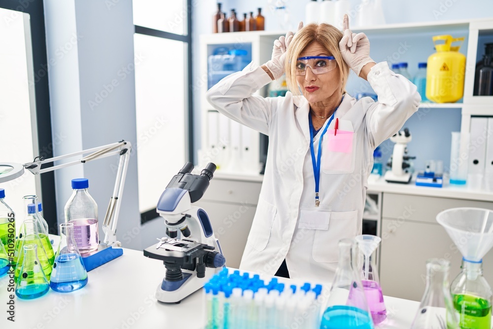 Middle age blonde woman working at scientist laboratory doing funny gesture with finger over head as bull horns Middle age blonde woman working at scientist laboratory doing funny gesture with finger over head as bull horns