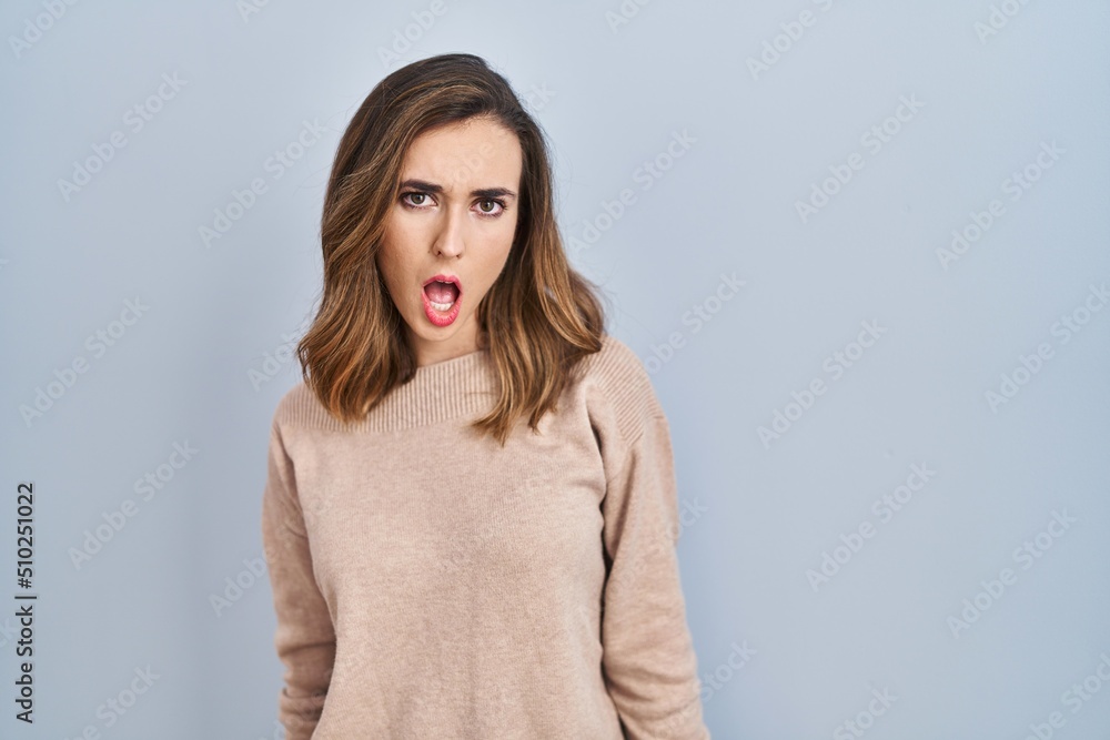 Young woman standing over isolated background in shock face, looking skeptical and sarcastic, surprised with open mouth