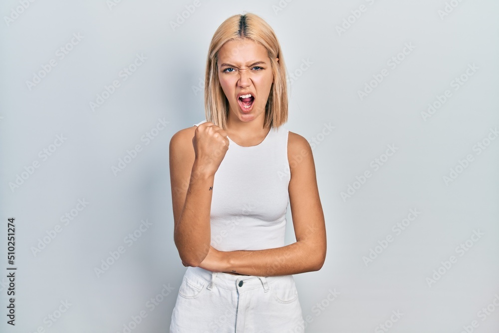 Beautiful blonde woman wearing casual style with sleeveless shirt angry and mad raising fist frustrated and furious while shouting with anger. rage and aggressive concept. Beautiful blonde woman wearing casual style with sleeveless shirt angry and mad raising fist frustrated and furious while shouting with anger. rage and aggressive concept.