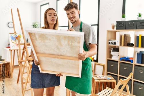 Young hispanic artist couple smiling happy holding canvas at art studio.