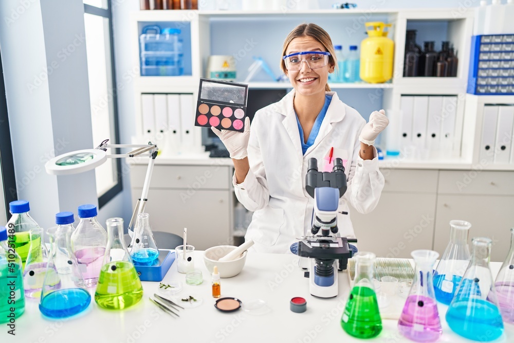 Young blonde woman working at scientist laboratory with make up screaming proud, celebrating victory and success very excited with raised arm Young blonde woman working at scientist laboratory with make up screaming proud, celebrating victory and success very excited with raised arm
