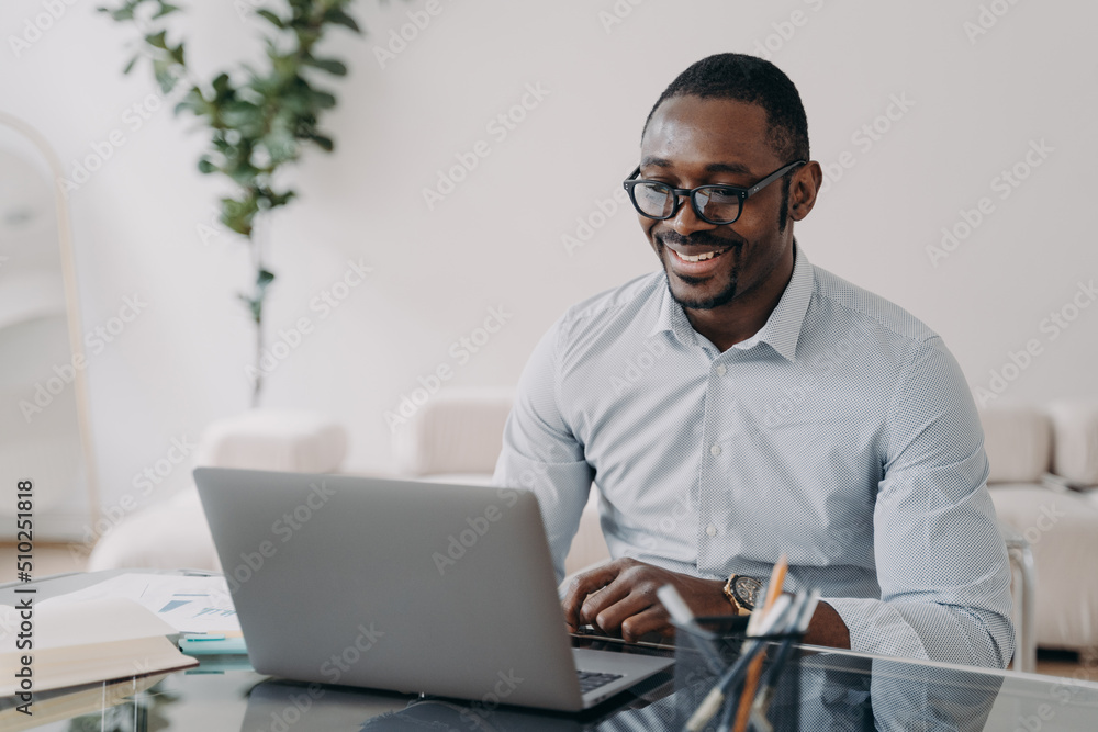 Smiling african american man wearing glasses working on business project at laptop at office desk Smiling african american man wearing glasses working on business project at laptop at office desk