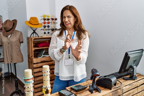 Middle age hispanic woman working as manager at retail boutique disgusted expression, displeased and fearful doing disgust face because aversion reaction. with hands raised