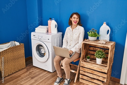 Young caucasian woman using laptop waiting for washing machine at laundry room