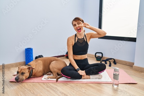 Young beautiful woman sitting on yoga mat doing peace symbol with fingers over face, smiling cheerful showing victory