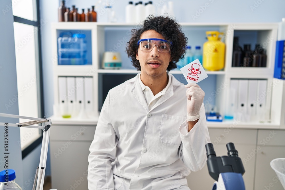 Hispanic man with curly hair working at scientist laboratory holding toxic banner scared and amazed with open mouth for surprise, disbelief face