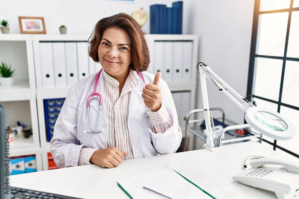Middle age hispanic woman wearing doctor uniform and stethoscope at the clinic doing happy thumbs up gesture with hand. approving expression looking at the camera showing success. Middle age hispanic woman wearing doctor uniform and stethoscope at the clinic doing happy thumbs up gesture with hand. approving expression looking at the camera showing success.