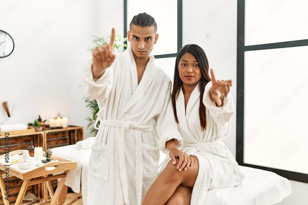 Young latin couple wearing towel standing at beauty center pointing with finger up and angry expression, showing no gesture Young latin couple wearing towel standing at beauty center pointing with finger up and angry expression, showing no gesture