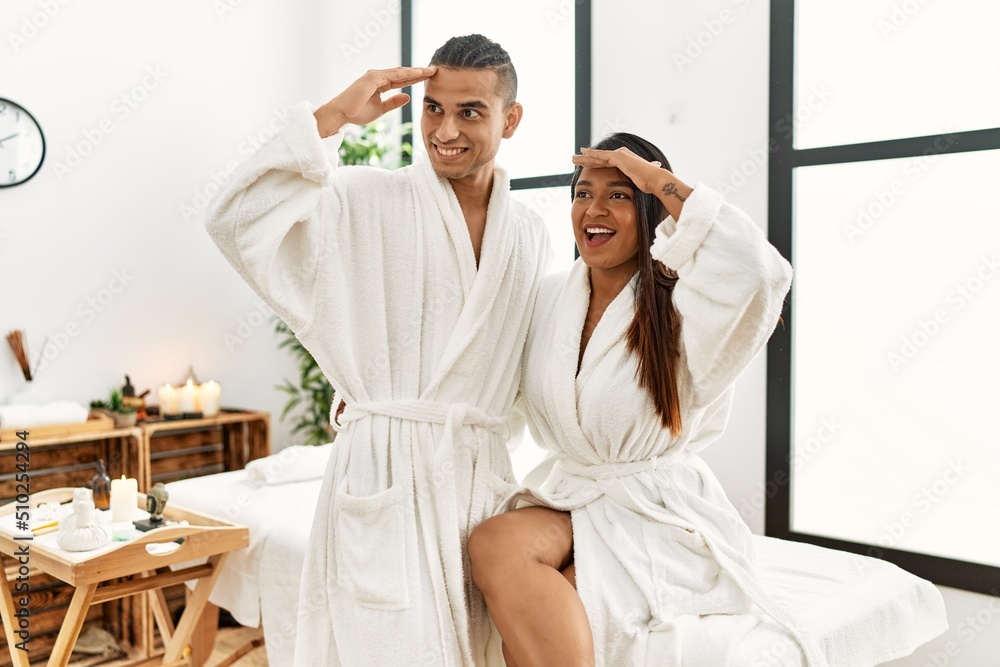 Young latin couple wearing towel standing at beauty center very happy and smiling looking far away with hand over head. searching concept. Young latin couple wearing towel standing at beauty center very happy and smiling looking far away with hand over head. searching concept.