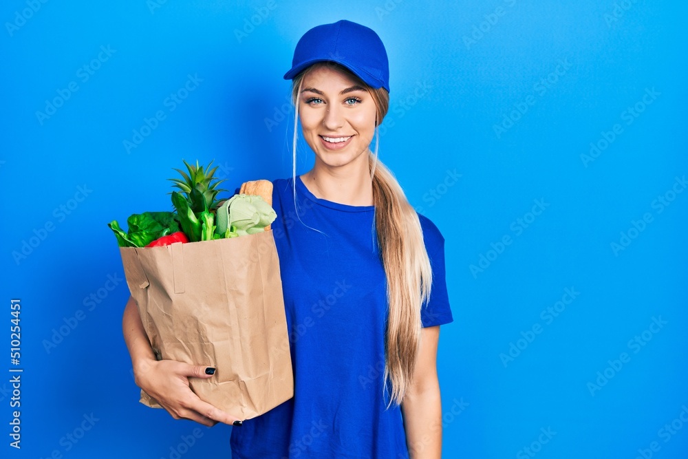 Young caucasian woman wearing courier uniform with groceries from supermarket with a happy and cool smile on face. lucky person. Young caucasian woman wearing courier uniform with groceries from supermarket with a happy and cool smile on face. lucky person.