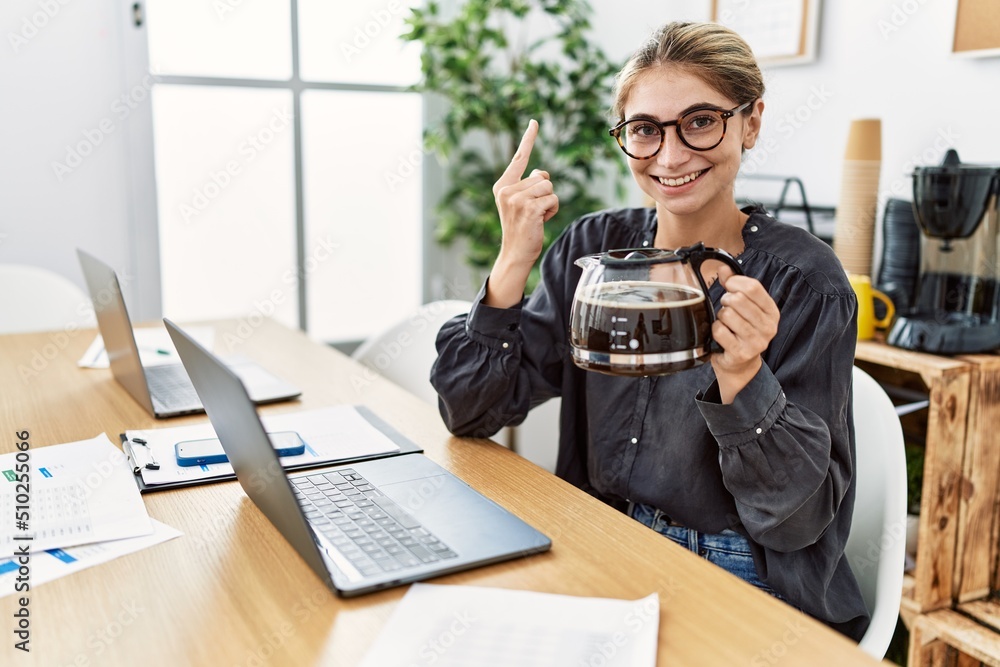 Young blonde woman working at the office holding coffee pot smiling with an idea or question pointing finger with happy face, number one Young blonde woman working at the office holding coffee pot smiling with an idea or question pointing finger with happy face, number one