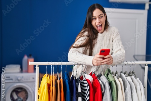 Young brunette woman waiting for laundry using smartphone celebrating crazy and amazed for success with open eyes screaming excited.
