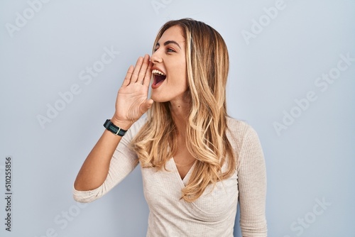 Young blonde woman standing over isolated background shouting and screaming loud to side with hand on mouth. communication concept.