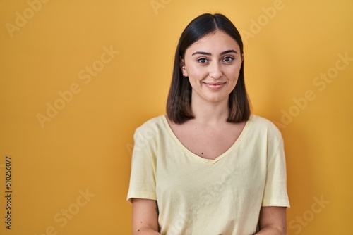 Hispanic girl wearing casual t shirt over yellow background with hands together and crossed fingers smiling relaxed and cheerful. success and optimistic